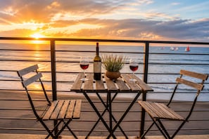 A wooden table with two chairs, a bottle of wine, and a glass of wine, set against a sunset view with sailboats on the water.