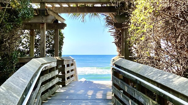 Ein hölzerner Steg führt zu einem Strand, darüber spendet eine Holzpergola Schatten.