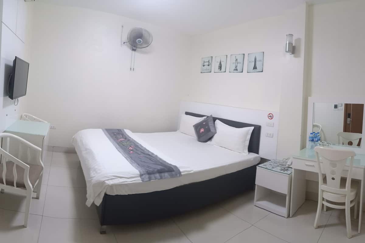 A bedroom with a bed, bedside table, chair, desk, and a wall-mounted fan.