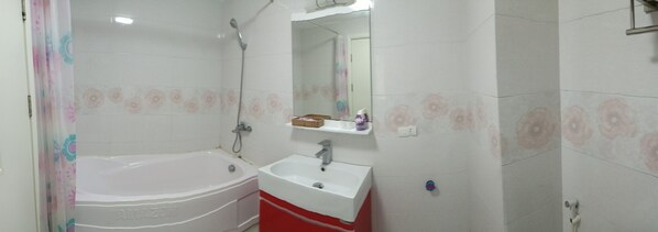 A bathroom with a white bathtub, a white sink, and a shower. There is a mirror above the sink and a shelf with a basket and a bottle. - Linh Anh Hotel (Hanoi)
