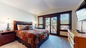 A bedroom with a large bed, a wooden nightstand, a desk with a computer, and a view of mountains through the windows.