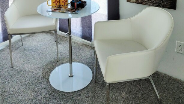 Una habitación moderna con una mesa de vidrio, dos sillas blancas, un adorno en la pared y vistas amplias a través de grandes ventanales.