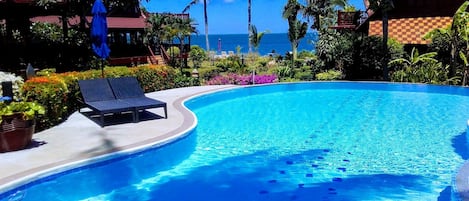 Una piscina azul cristalina con tumbonas, rodeada de plantas tropicales y palmeras, con vista al océano.