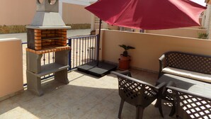 Une terrasse avec un barbecue, une table avec des chaises, une plante en pot et un parasol rouge.