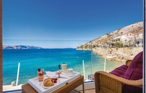 Un balcon avec une table dressée pour le petit-déjeuner, offrant une vue imprenable sur la côte, avec une eau d’un bleu limpide et un paysage montagneux en arrière-plan.