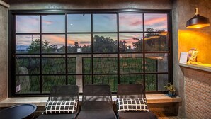 A room with a large window offering a view of a sunset over a green landscape. There are two lounge chairs with patterned cushions. - Be Live Residence (Thep Krasattri)