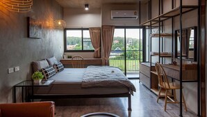 A modern bedroom with a bed, bedside tables, a desk, and a chair. There is a large window with curtains and a view of greenery outside. - Be Live Residence (Thep Krasattri)