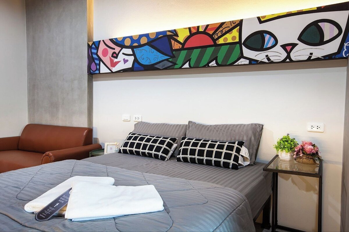 A hotel room with a bed, a brown leather chair, a small table with a vase of flowers, and a colorful abstract painting above the bed.
