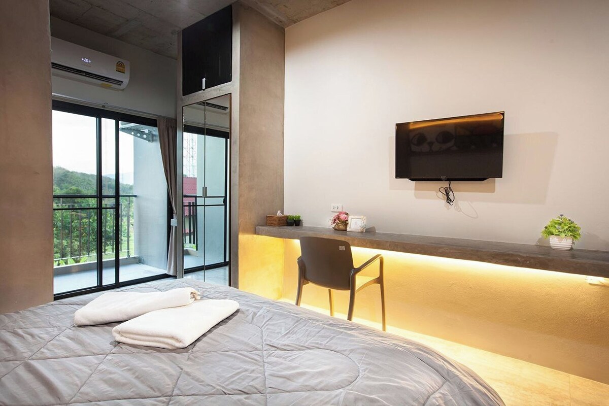 Quarto de hotel moderno com uma cama grande, uma escrivaninha com cadeira, televisão de tela plana na parede e uma varanda com vista para o verde.