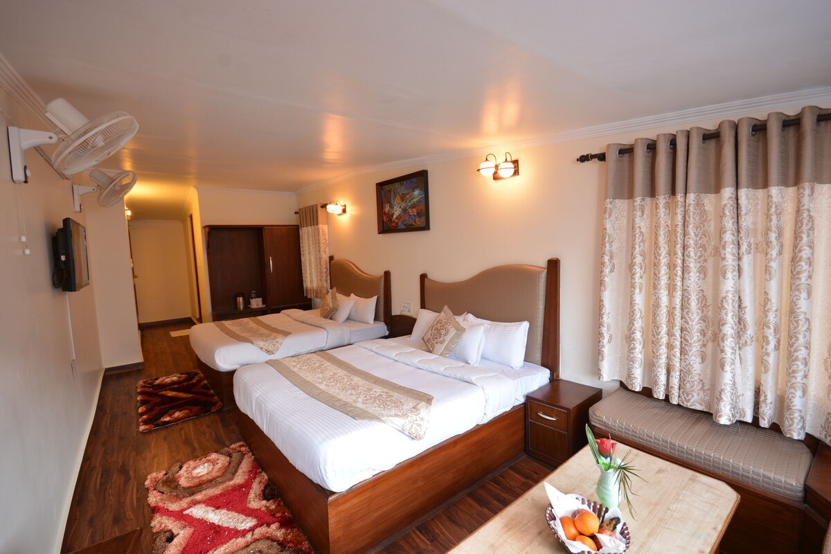 A double bed with a wooden headboard, a bedside table, a small table with fruit and a vase, a standing fan, and a window with curtains.