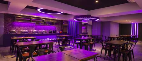 Un restaurante con mesas y sillas de madera, una zona de bar con pared de piedra e iluminación ambiental morada.
