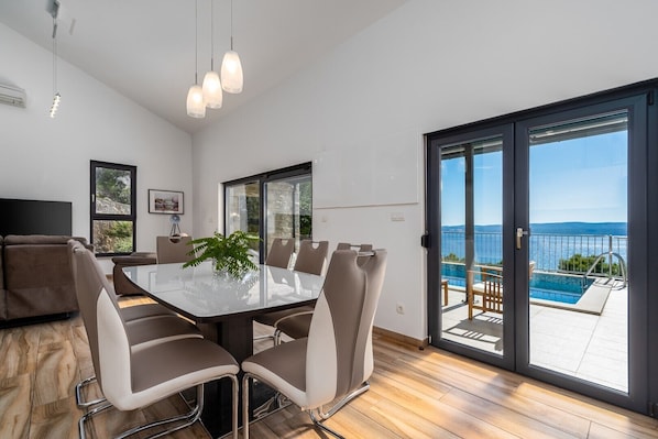 Un comedor moderno con una mesa y sillas de vidrio, una planta en maceta y vistas a una piscina y montañas a través de un ventanal corredera.