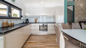 Eine moderne Küche mit weißen Schränken, einer schwarzen Arbeitsplatte und einem eingebauten Backofen. Ein Essbereich mit Tisch und Stühlen sowie ein Fenster mit Blick ins Freie.