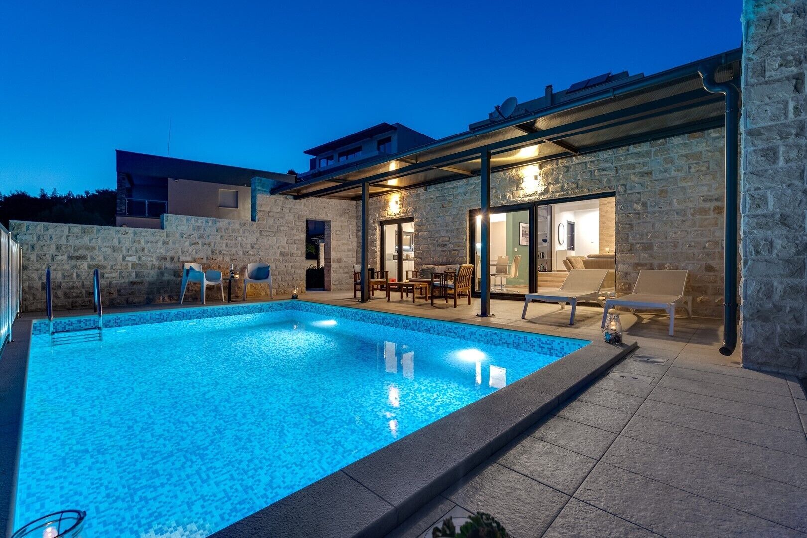 A swimming pool with a stone wall and a covered patio area with chairs and a table.