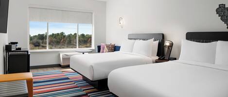 A hotel room with two beds, a large window with a view of trees, a colorful striped rug, and a small table with a lamp.