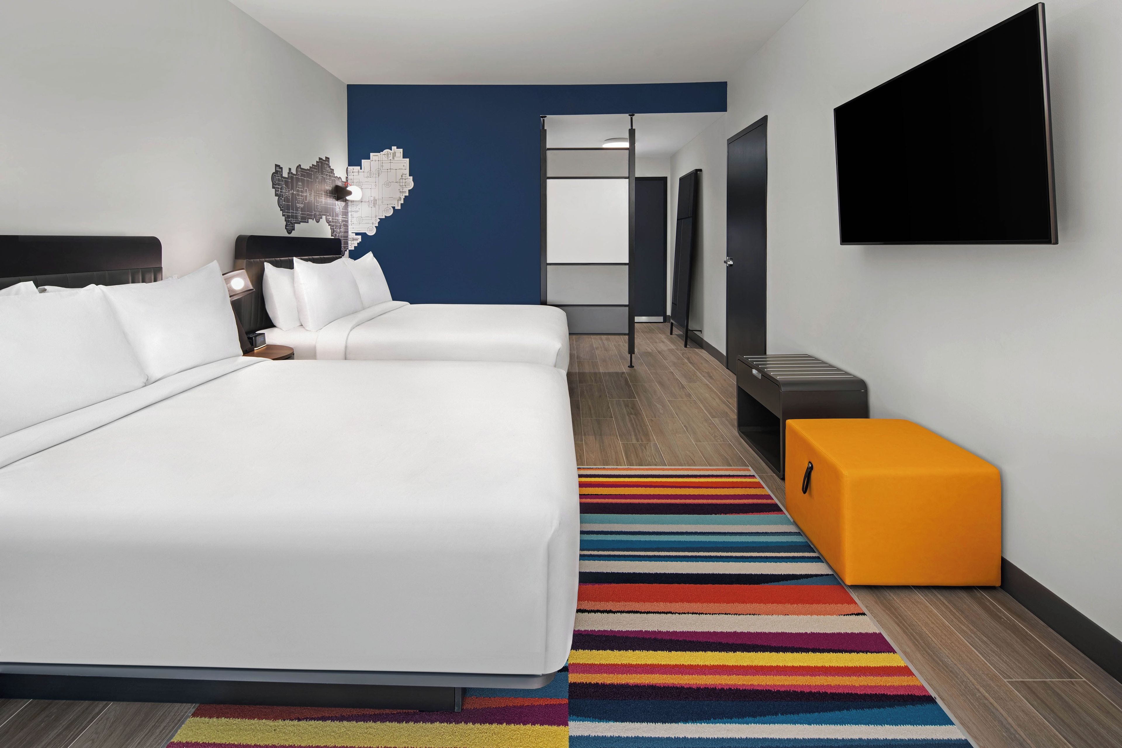 Ein Hotelzimmer mit einem Bett, einem Fernseher, einem kleinen orangefarbenen Hocker und einem farbenfrohen StreifenTeppich.
