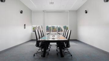 Una sala de conferències amb una taula llarga, cadires, un projecte i una finestra que dona a vistes d'arbres.