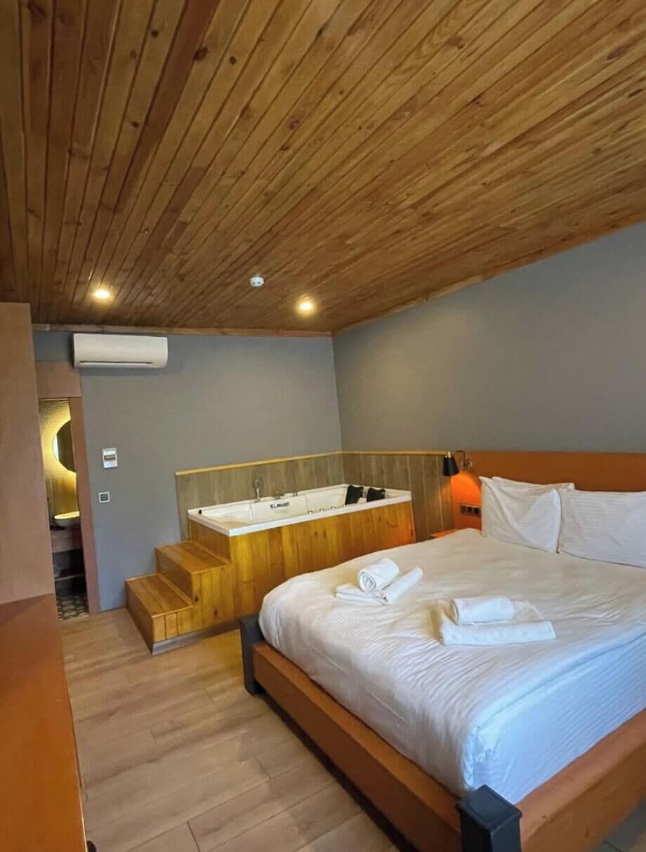 Een slaapkamer met een houten plafond, een groot bed, een nachtkastje van hout, een ingebouwd bad en een spiegel boven de wastafel.