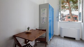 A small room with a wooden table and chairs, a blue locker, and a window with a view of buildings and trees. - Ostello Corniglia (Vernazza)