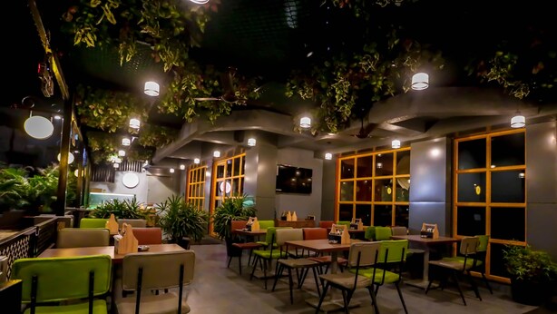 An indoor dining area with tables and chairs, surrounded by large windows and potted plants. - Hotel Imperial (Vasai)