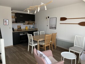 Une cuisine compacte dotée d’appareils électroménagers modernes, un coin repas avec une table en bois et des chaises blanches, et un coin salon confortable avec un fauteuil rose et une petite table blanche.