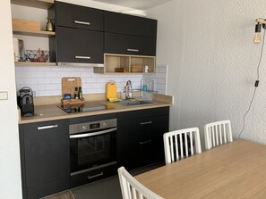 Una cocina compacta con armarios negros, horno integrado, lavamanos y mesa de comedor de madera con sillas blancas.