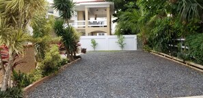 A building with a white facade, a balcony, and a gravel driveway surrounded by various plants.