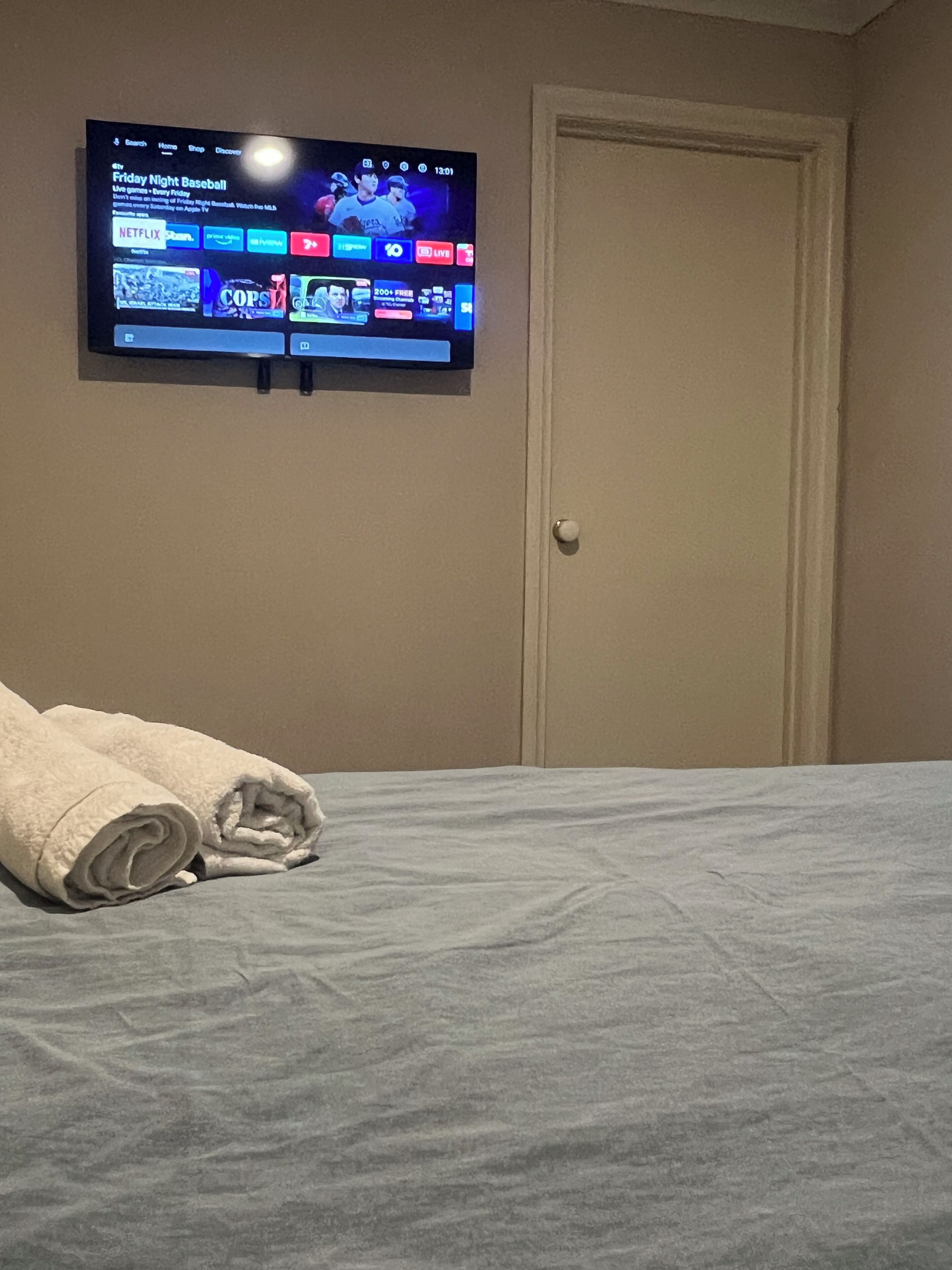 Hotellihuone, jossa on sänky, seinälle asennettu televisio, jolta näkyy urheilukanava, ja ovi.