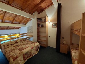 Quarto com cama beliche de madeira, uma cama de solteiro com colcha, mesa de cabeceira de madeira, prateleira com livros e luminária na parede.