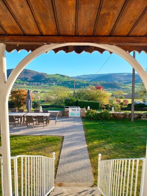 A covered patio with a gravel path leading to a white fence, overlooking a green landscape with hills and a clear sky.