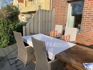 Een terras buiten met een tafel en rieten stoelen, een houten bank en een bakstenen muur.