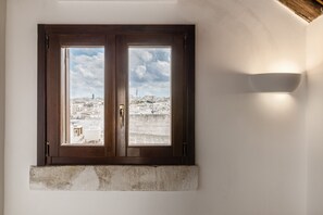 A room with a large window overlooking a cityscape, a stone ledge below the window, and a wall-mounted light fixture. - Le Cupole - Suite Panoramica by Wonderful Italy (Ostuni)
