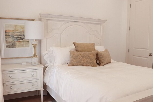 Un lit bien fait, avec du linge de lit blanc et deux oreillers marron, une table de chevet blanche avec une lampe, et un tableau encadré au mur.