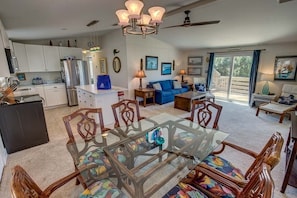 A spacious living area with a glass-top dining table, a blue sofa, and a kitchen with white cabinets and a refrigerator.