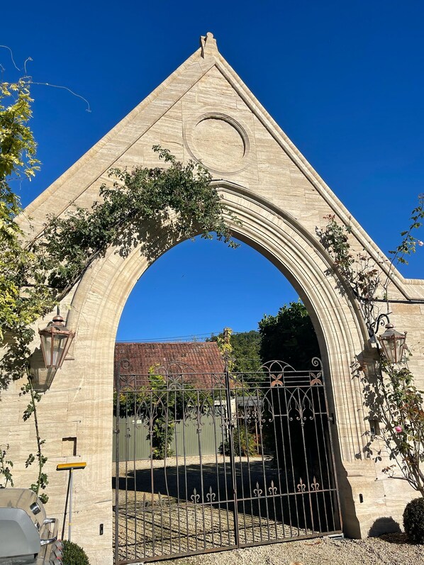 Une arche en pierre surmontée d’un portail en métal, entourée de verdure et s’ouvrant sur un ciel bleu clair.