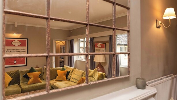 A hotel lobby with a large mirror reflecting a green sofa with yellow cushions, a white fireplace, and a wall-mounted lamp. - Spacious Country Cottage Sleeps 7 Near Bath set in a Country House Estate (Bradford-on-Avon)