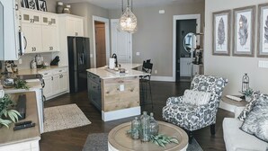 Une cuisine moderne avec des meubles blancs, un îlot central et un coin repas. On y trouve un fauteuil à motifs et une table basse ronde agrémentée d’objets décoratifs.