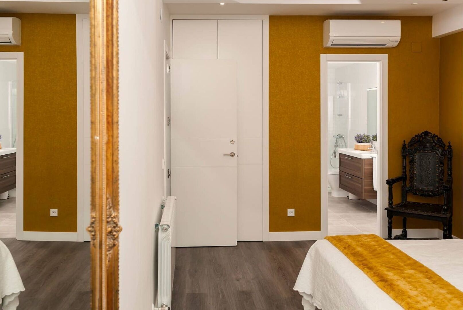 Une chambre avec une porte blanche, une chaise en bois et un lit recouvert d’une couvre-lit blanche et dorée.