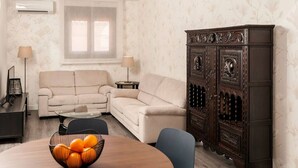 Ein Wohnzimmer mit einem beigefarbenen Sofa, einem dunklen Holzschrank, einem runden Tisch mit Obstschale und zwei Stehlampen.