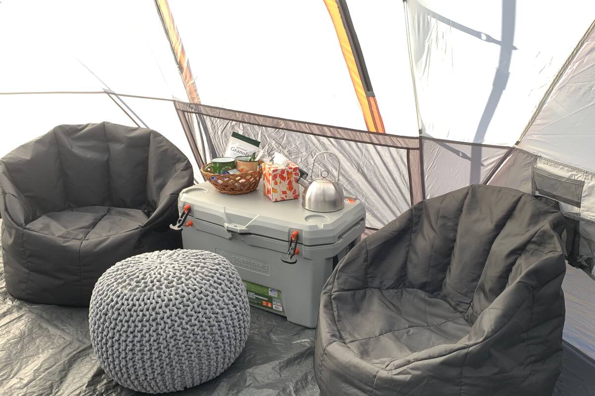A tent with two grey bean bag chairs, a small round ottoman, and a cooler with items like a kettle and granola.