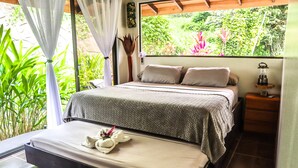 A bedroom with a bed, bedside table, and a window with a view of greenery.