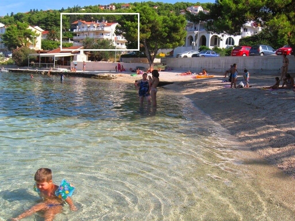 A beach scene with clear water, people swimming and sunbathing, and buildings in the background.