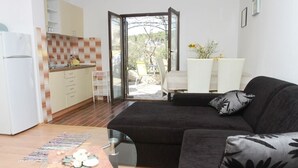 Ein modernes Wohnzimmer mit einem schwarzen Sofa, einem Glastisch und einer Küchenzeile, inklusive eines weißen Kühlschranks und beiger Schränke.