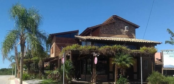 A two-story building with a brick facade, a tiled roof, and a covered porch area with plants. - Residencial Banzaê (Garopaba)