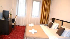 Quarto de hotel com uma cama, uma escrivaninha com televisão e um tapete vermelho.