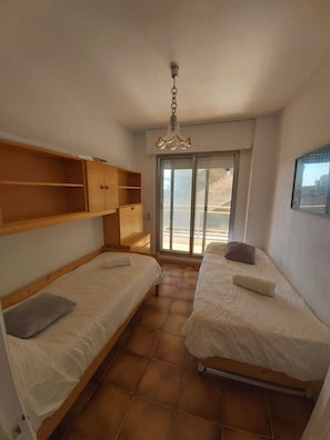 A small, compact room with two single beds, a wooden wardrobe, a ceiling light, and a sliding glass door leading to an outdoor area.