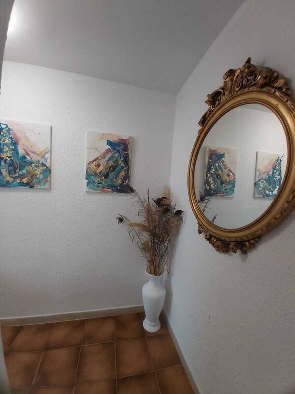 Un pasillo con dos cuadros abstractos en la pared, un jarrón blanco con plantas secas y un espejo con marco dorado.