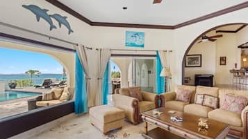 Ein großzügiger Wohnbereich mit einem großen Fenster mit Blick auf einen Pool und das Meer, ausgestattet mit einer Couch, Sesseln und einem Couchtisch.