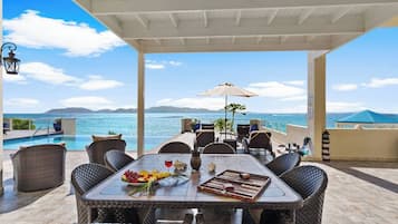Ein überdachter Essbereich im Freien mit Korbmöbeln, einem Tisch mit Früchten und einem Brettspiel, mit Blick auf einen Pool und das Meer.