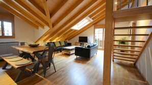 Un loft espacioso con techos altos, vigas de madera, una zona de comedor con una mesa larga y bancos, un sofá y una escalera.